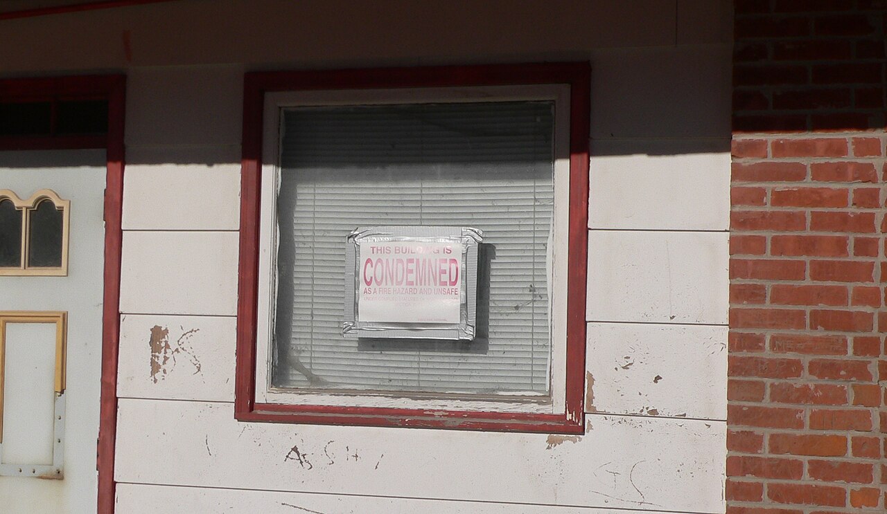 A CONDEMNED notice taped inside the window of a weathered building with closed blinds.
