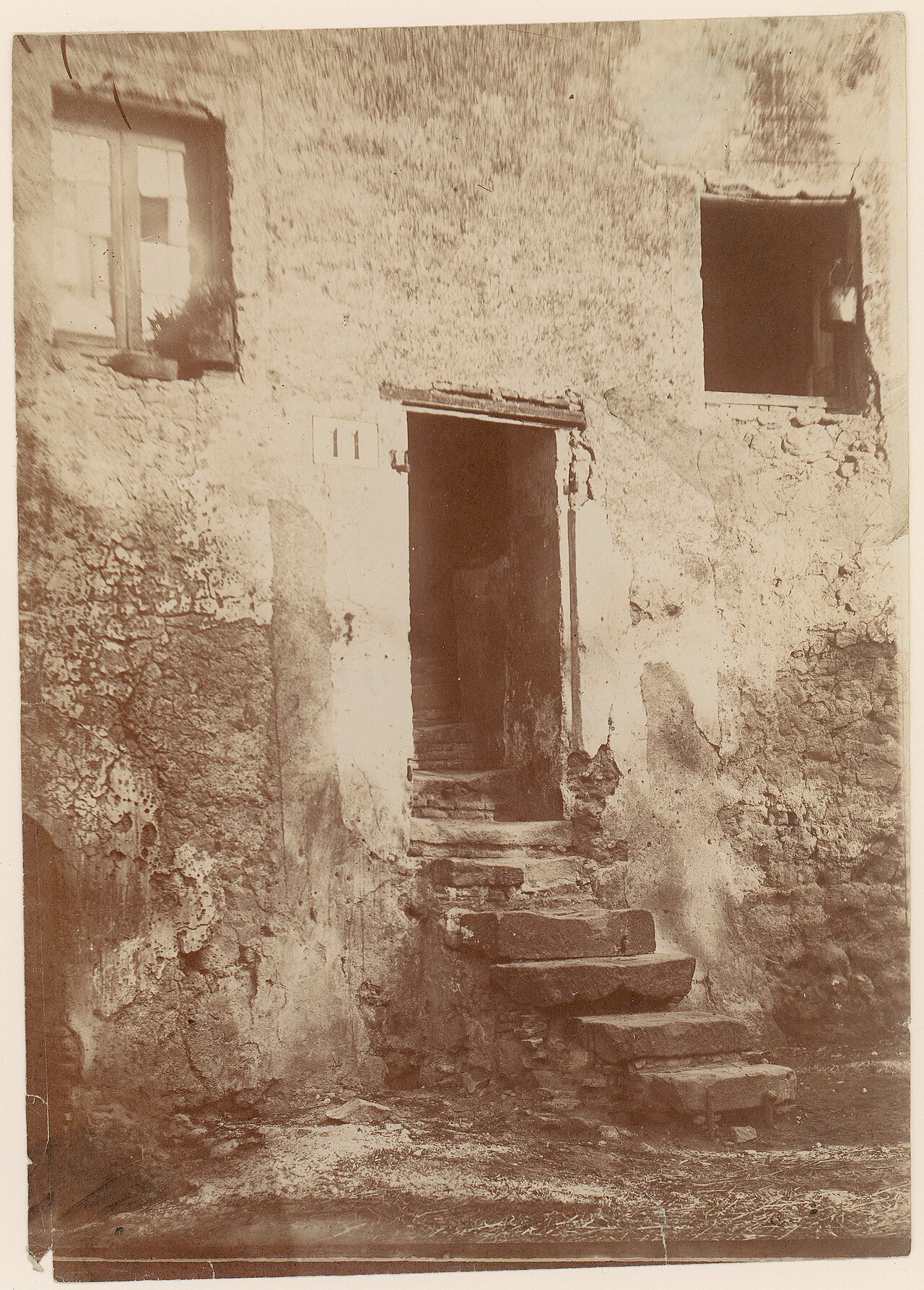 A sepia photograph of a crumbling plastered house with a dark open doorway above worn stone steps.