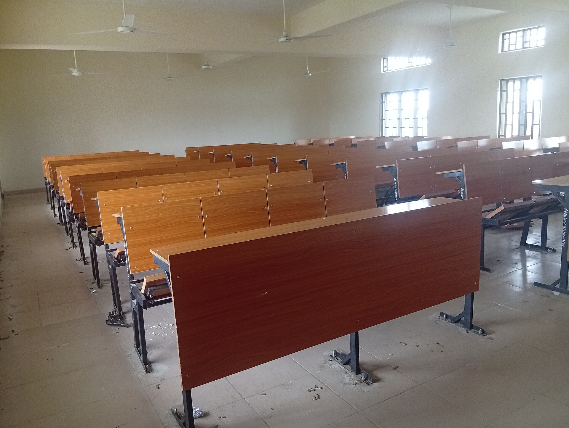 Rows of empty wooden lecture benches in a bare classroom with ceiling fans.