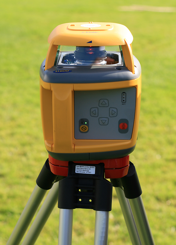 A yellow rotary construction laser on a tripod in a grassy field.