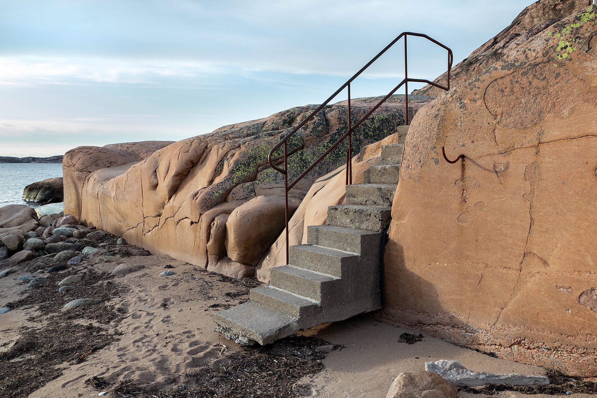Concrete steps with a metal handrail climbing a smooth granite rock face on a beach and stopping partway up.